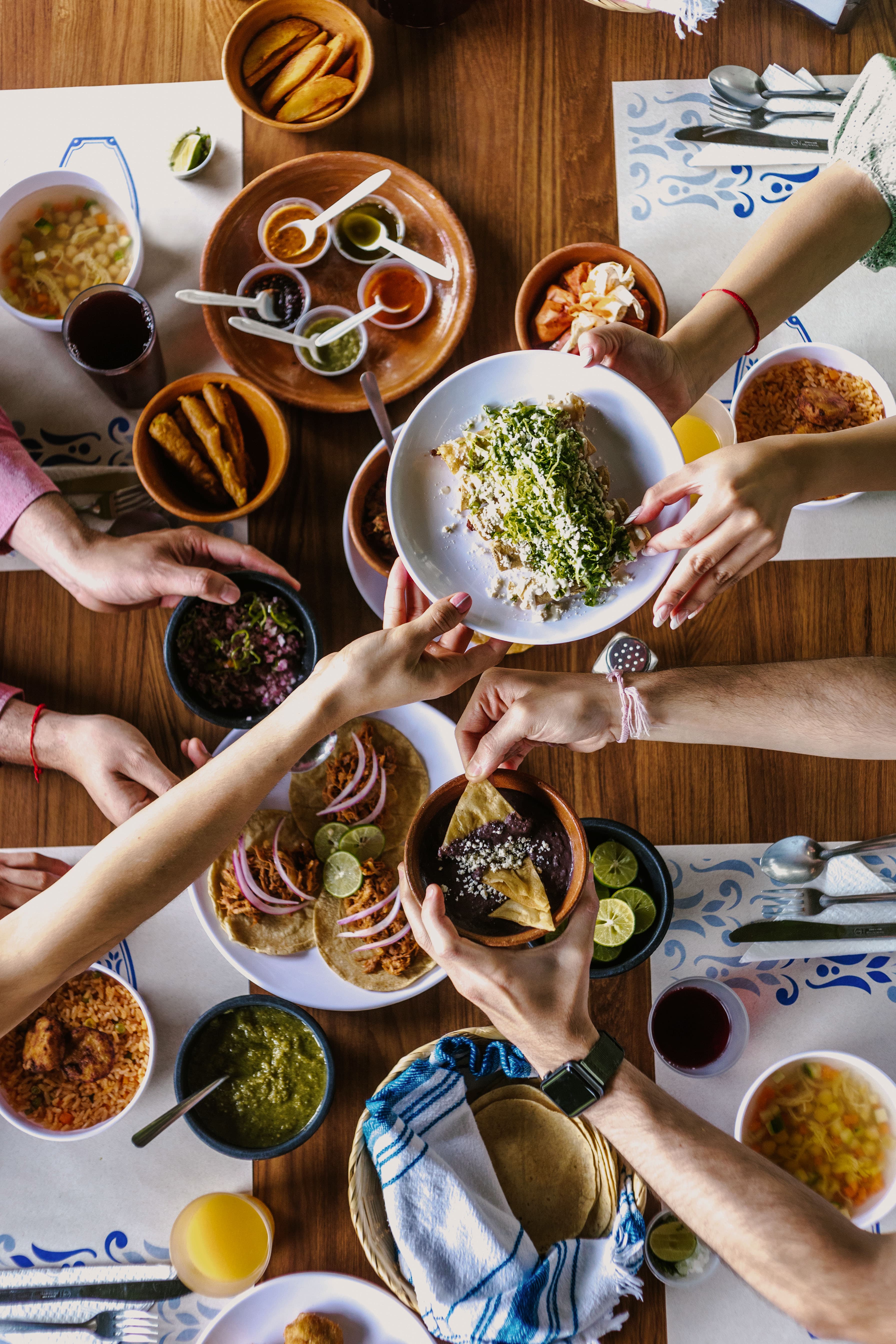 Hands reaching across a wooden table, passing bowls and plates of food between them.